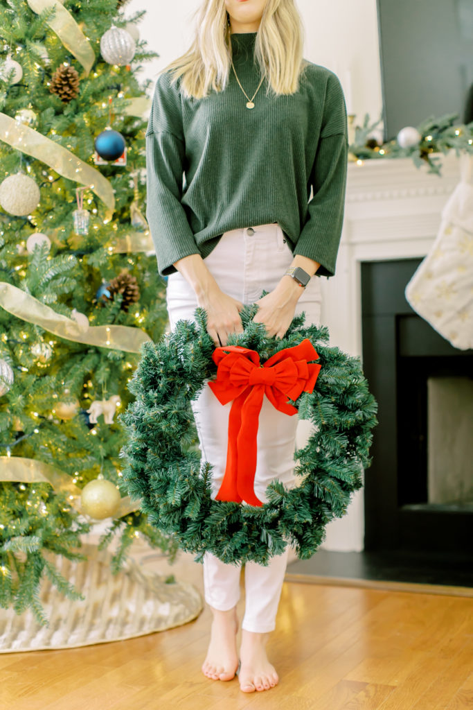Holding wreath with red bow in front of christmas tree and mantle