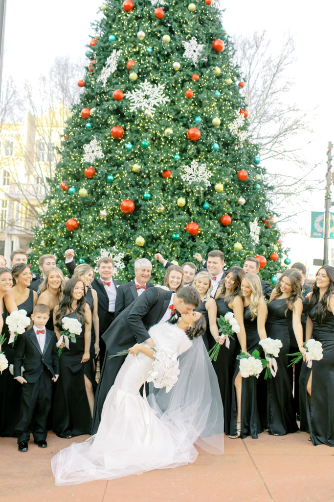 bride and groom dip kiss in the middle of their bridal party in front of the christmas tree | Photo by Mary Catherine Echols