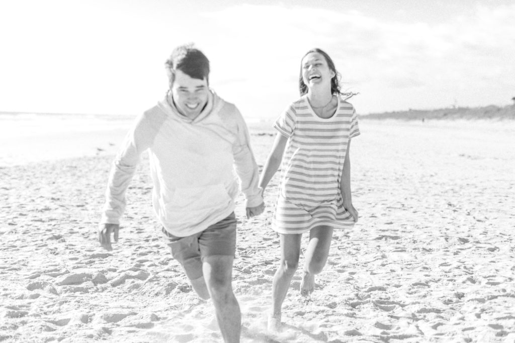black and white photo of engaged couple holing hands and running on beach